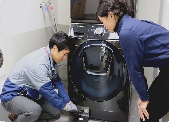삼성전자서비스 수리 엔지니어가 출장 점검 서비스를 신청한 고객에게 플러스 원 케어 서비스의 일환으로 드럼세탁기 배수펌프 잔수 제거 방법을 설명하고 있는 모습